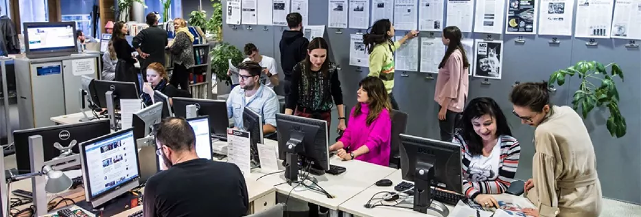 People in a newsroom. They are writing, looking on screens and talking about papers.