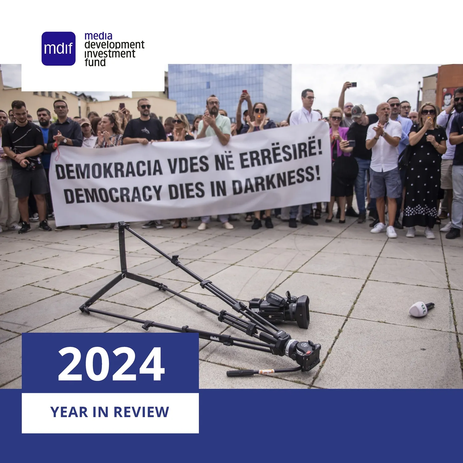 A group of protesters stands in a public square holding a large bilingual banner that reads "DEMOKRACIA VDES NË ERRËSIRË! DEMOCRACY DIES IN DARKNESS!" in bold black letters. The crowd is diverse, with many people clapping or recording on their phones. In the foreground, a toppled video camera, tripod, and microphone lie abandoned on the pavement, symbolizing a silenced press. Overlaid on top wth MDIF logo and in the bottom left corner is a graphic with bold text reading "2024 YEAR IN REVIEW" in white and blue.