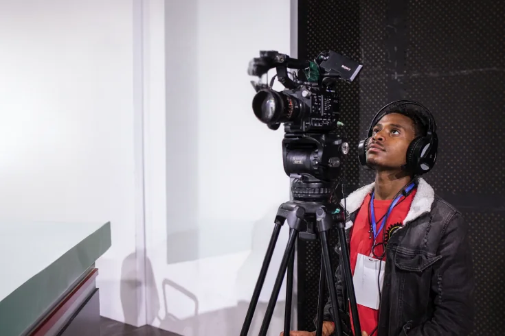 Young man wearing headphones operates a professional video camera on a tripod, focusing intently inside a studio setting