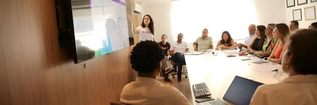 A woman giving a presentation to a group of people seated around a large conference table, with a screen displaying slides on the wall.