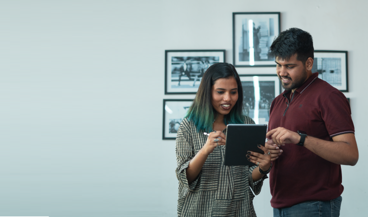 Two media workers standing indoors and looking at a tablet together, smiling while discussing something on the screen, with framed black-and-white photographs on the wall in the background.