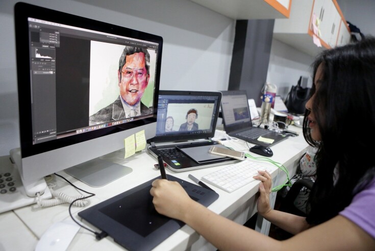 A woman works at a desk using a digital drawing tablet, creating illustrations on a computer. On the large monitor is a digital portrait of a man in a suit, while the laptop next to it displays a cartoon-style drawing of two people. Multiple laptops, a smartphone, and sticky notes are on the desk