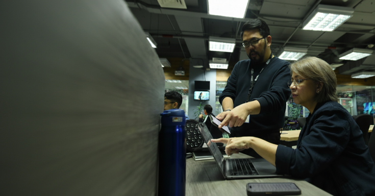 In an office setting with fluorescent lighting, a man stands next to a seated woman as they discuss something on her laptop. The man points at the screen while holding a phone, and the woman also gestures toward the laptop. Other people work in the background, with monitors and glass walls visible.