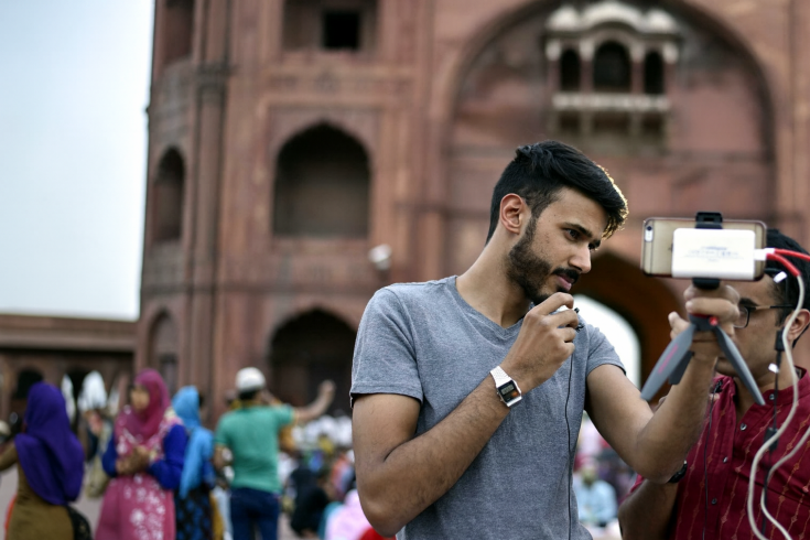 Man holding a microphone and speaking while being filmed on a smartphone mounted on a tripod, with a historic red stone building and a crowd of people in the background.