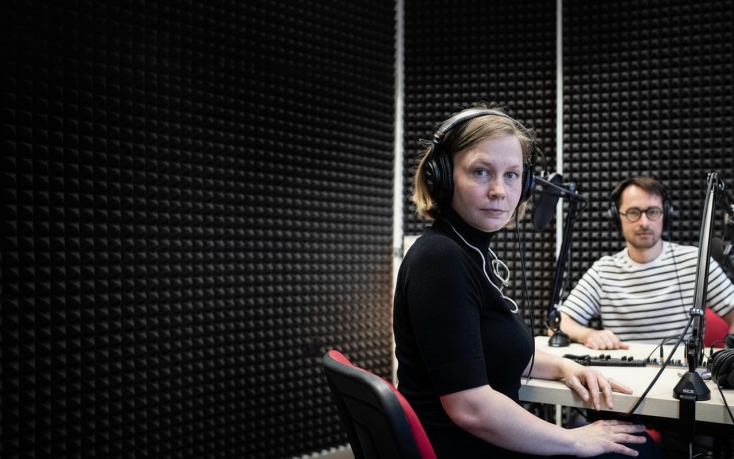 Two people in a recording studio sit at a table with microphones and headphones. The woman in the foreground, wearing a black top and large headphones, looks directly at the camera. Behind her, a man in glasses and a striped shirt sits at the table with audio equipment. The walls are covered with black acoustic foam panels.