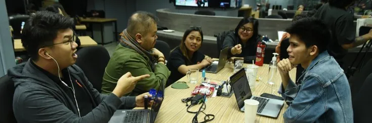 Group of people sitting around a table in a newsroom, engaged in an animated discussion with laptops, drinks, and snacks on the table